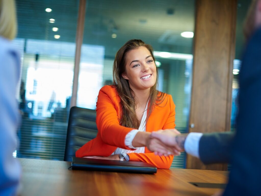 Woman in bright suit jacket shaking hands with man in meeting room.